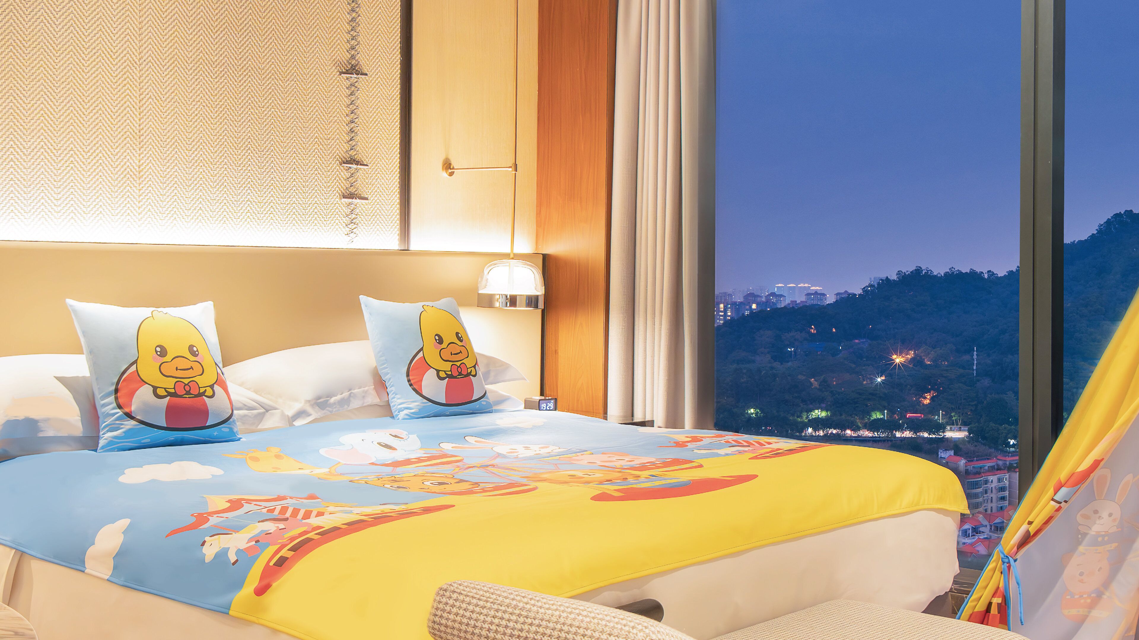 Habitación de hotel con una cama grande, dos almohadas con personajes de dibujos animados, una mesita de noche con lámpara y vistas a un paisaje urbano al anochecer.