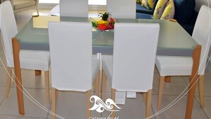 Un coin repas avec une table en verre et des chaises blanches, un saladier de fruits sur la table et une vue sur l’océan par une porte-fenêtre coulissante.