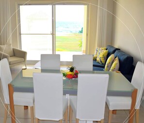 A dining area with a glass table and white chairs, a bowl of fruit on the table, and a view of the ocean through a sliding glass door.