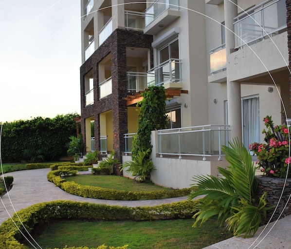 A multi-story building with balconies, a landscaped garden with hedges and plants, and a clear sky.