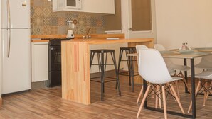 Cozinha compacta com geladeira, micro-ondas e bancada de madeira. Há uma área de refeições com mesa e cadeiras.