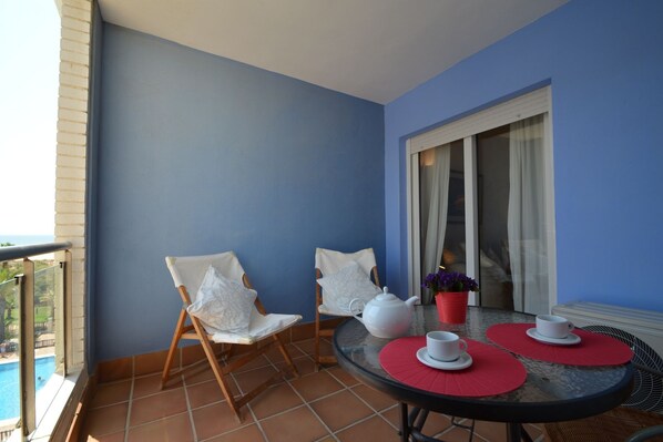Ein Balkon mit zwei weißen, gepolsterten Stühlen, einem runden Tisch mit roten Unterlagen, weißen Teekannen und einem Blumentopf.