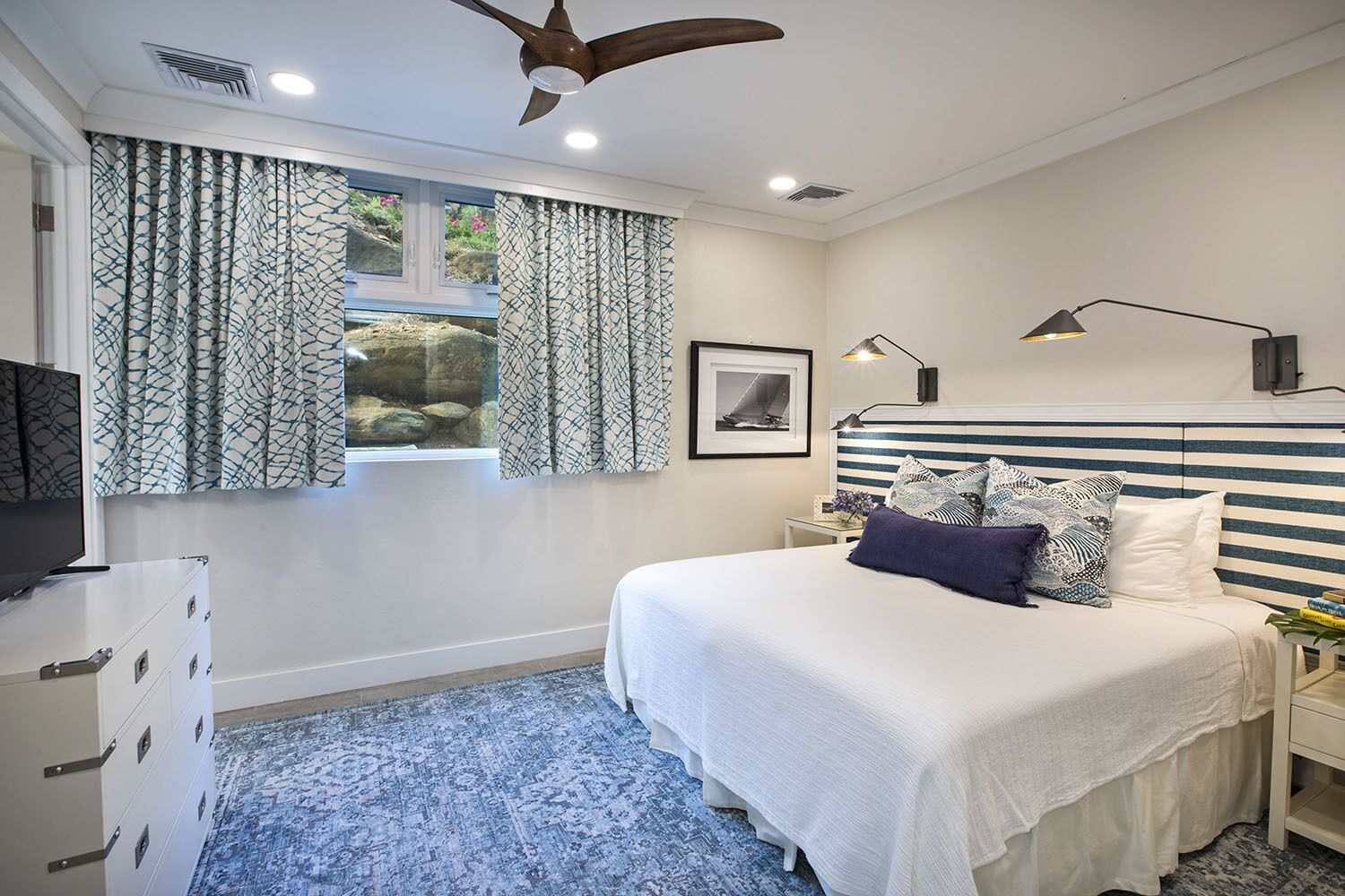 Un dormitorio con cama, ventana con cortinas, ventilador de techo, lámpara de noche y televisor sobre una cómoda.
