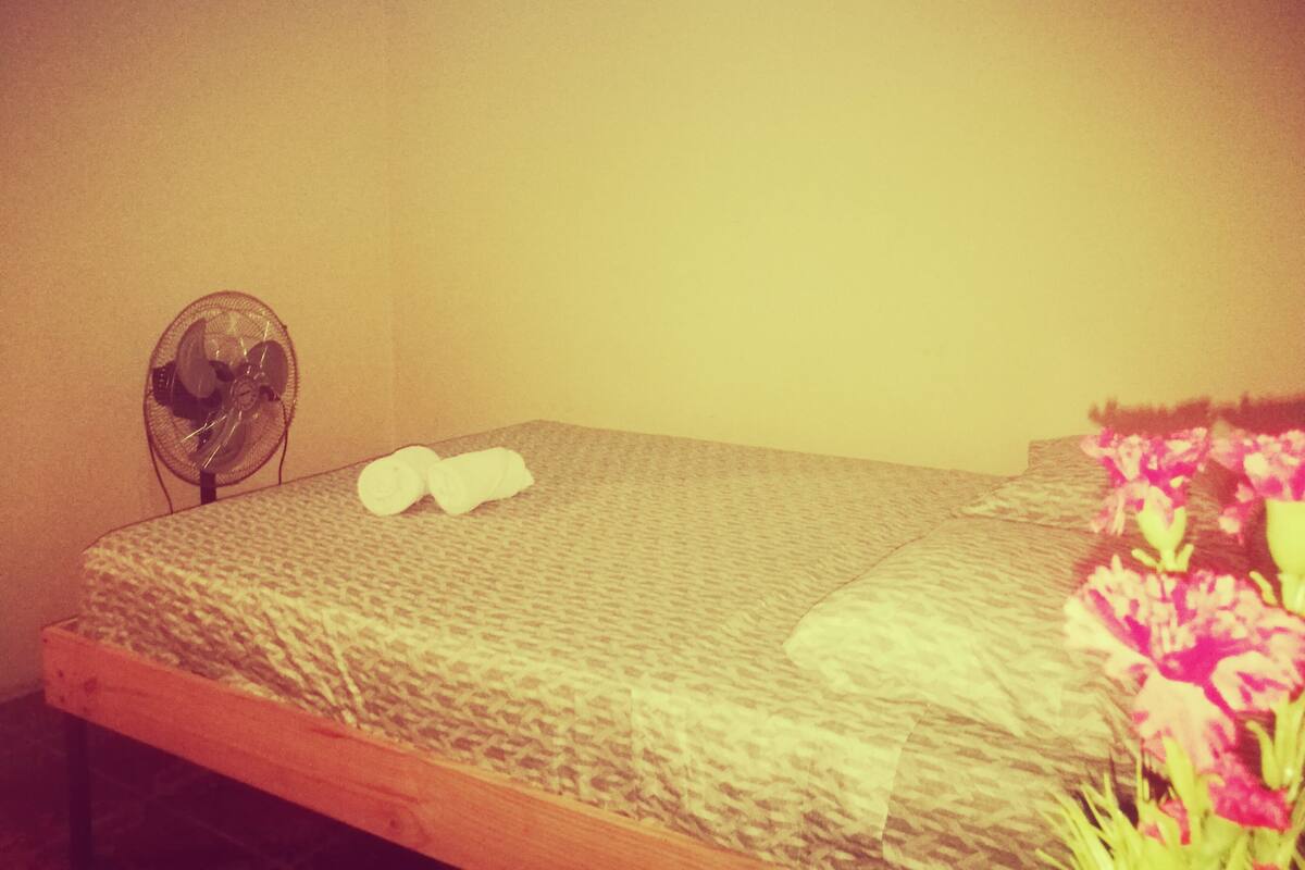 A bed with a patterned bedspread, a fan, and a pair of rolled towels.