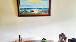 A dining area with a wooden table and chairs, a painting of a sunset over water, and a small potted plant on the table. - Guesthouse Morpho CR (Quepos)
