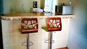 A kitchen with two bar stools, a microwave, a coffee maker, and a window with curtains. - Guesthouse Morpho CR (Quepos)