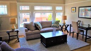 A living room with a sofa, coffee table, and two side tables with lamps. There are windows with a view of buildings outside.