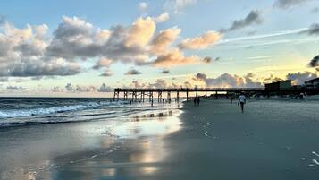 A beach with a pier extending into the ocean, people walking along the shore, and a clear sky with clouds.