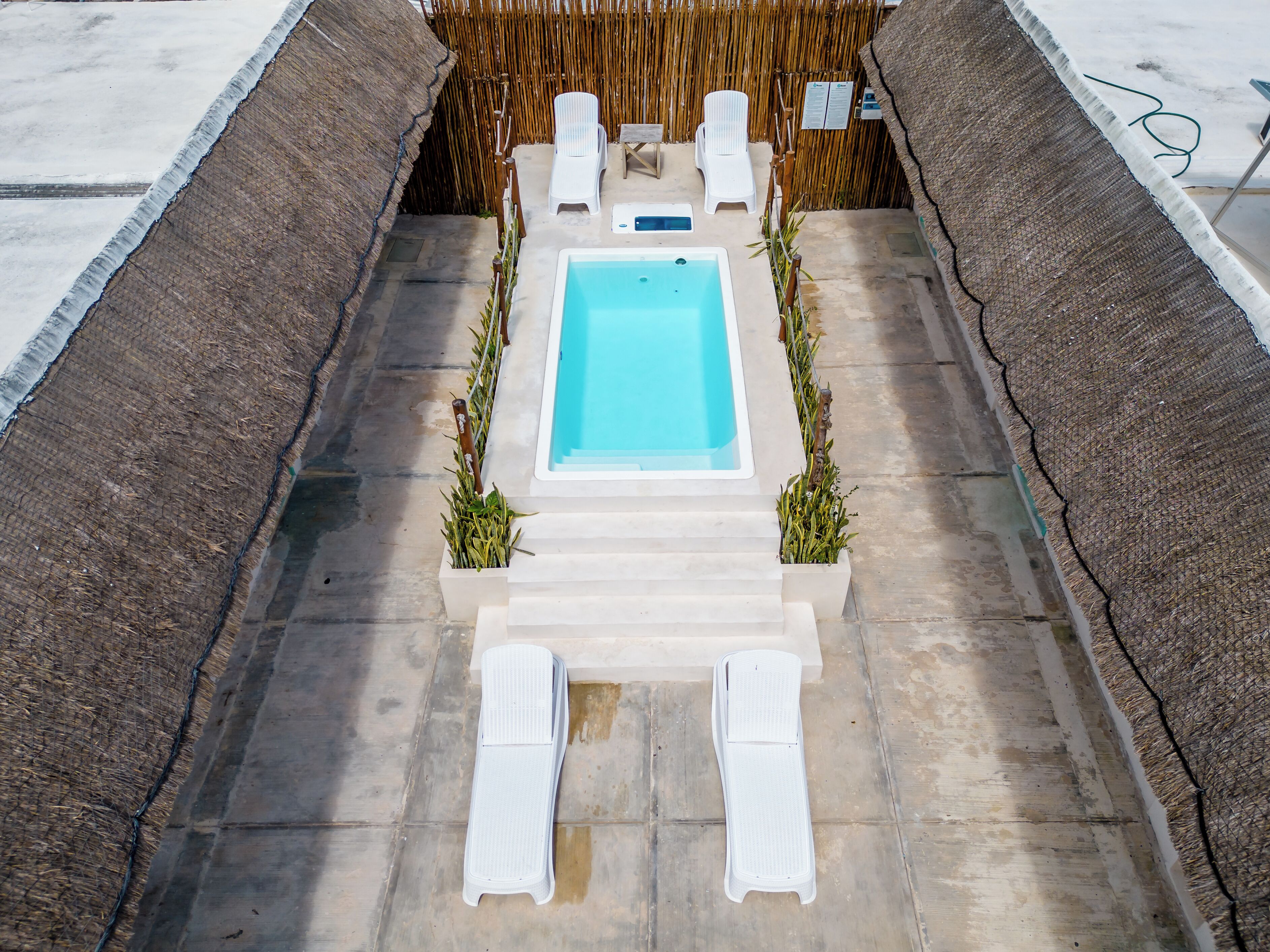 A rectangular pool with a white border, surrounded by a concrete deck and a bamboo fence. There are two white lounge chairs and a small table with a chair.