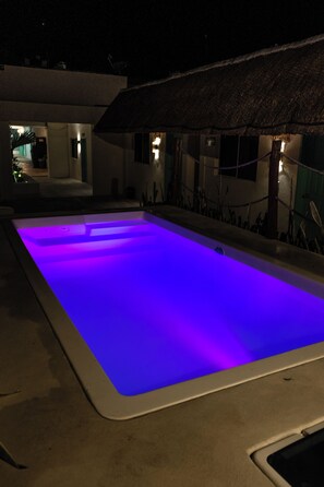 A swimming pool with blue lighting, surrounded by a building with a thatched roof and a small garden area. - Hotel Pelicano (Chiquilá)