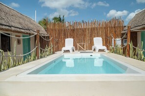 A swimming pool with two white lounge chairs, surrounded by a bamboo fence and thatched-roof huts. - Hotel Pelicano (Chiquilá)