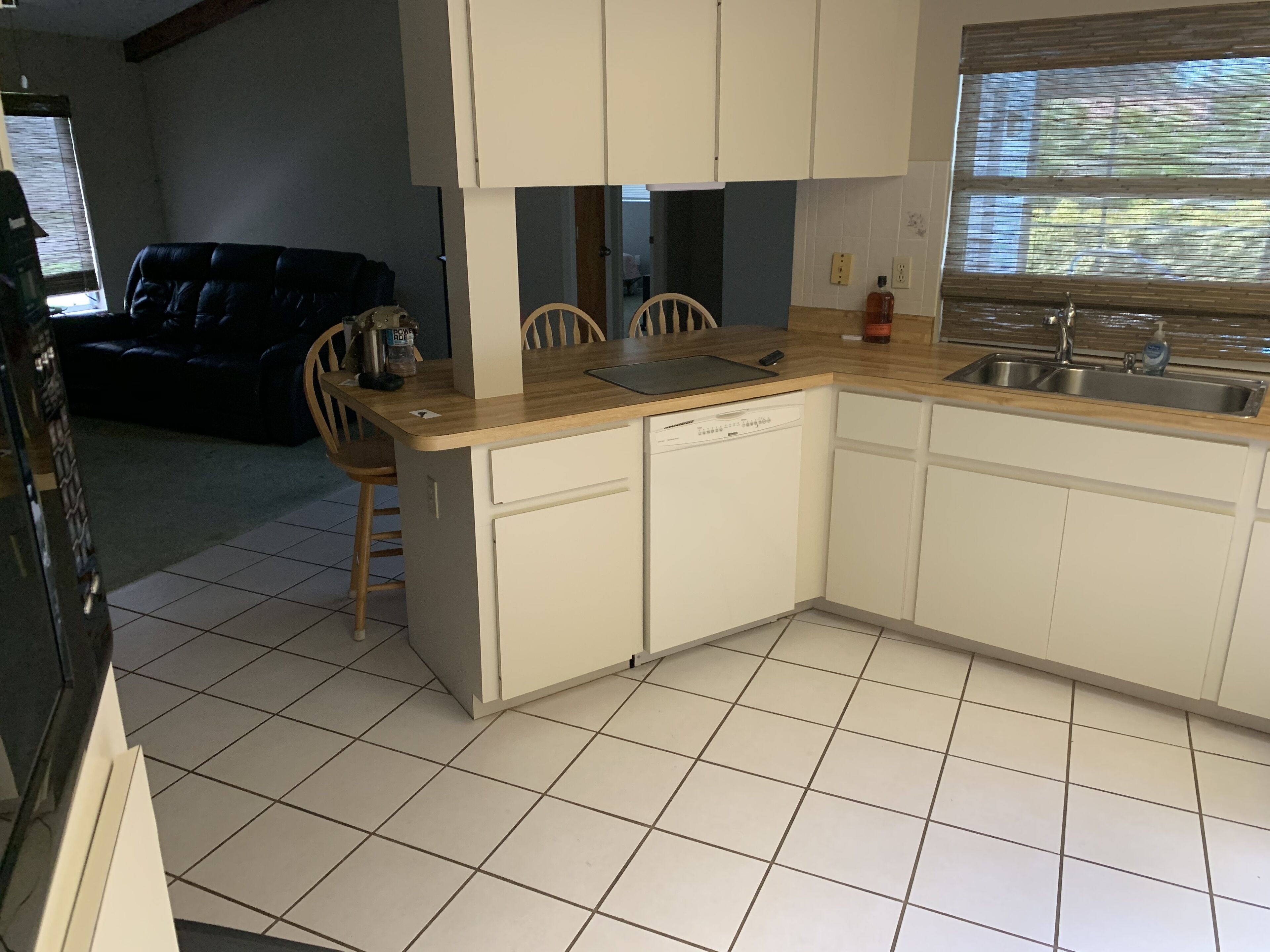 Una cocina compacta con armarios blancos, lavavajillas y un lavamanos. Hay una zona de comedor con un sofá negro y sillas de madera.