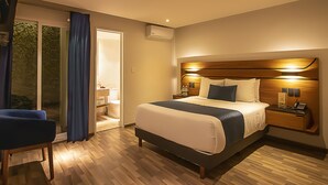 A modern hotel room with a large bed, a bedside table with a phone and a lamp, a chair, and a sliding glass door leading to a bathroom. - Hotel Boutique Maria (Morelia)