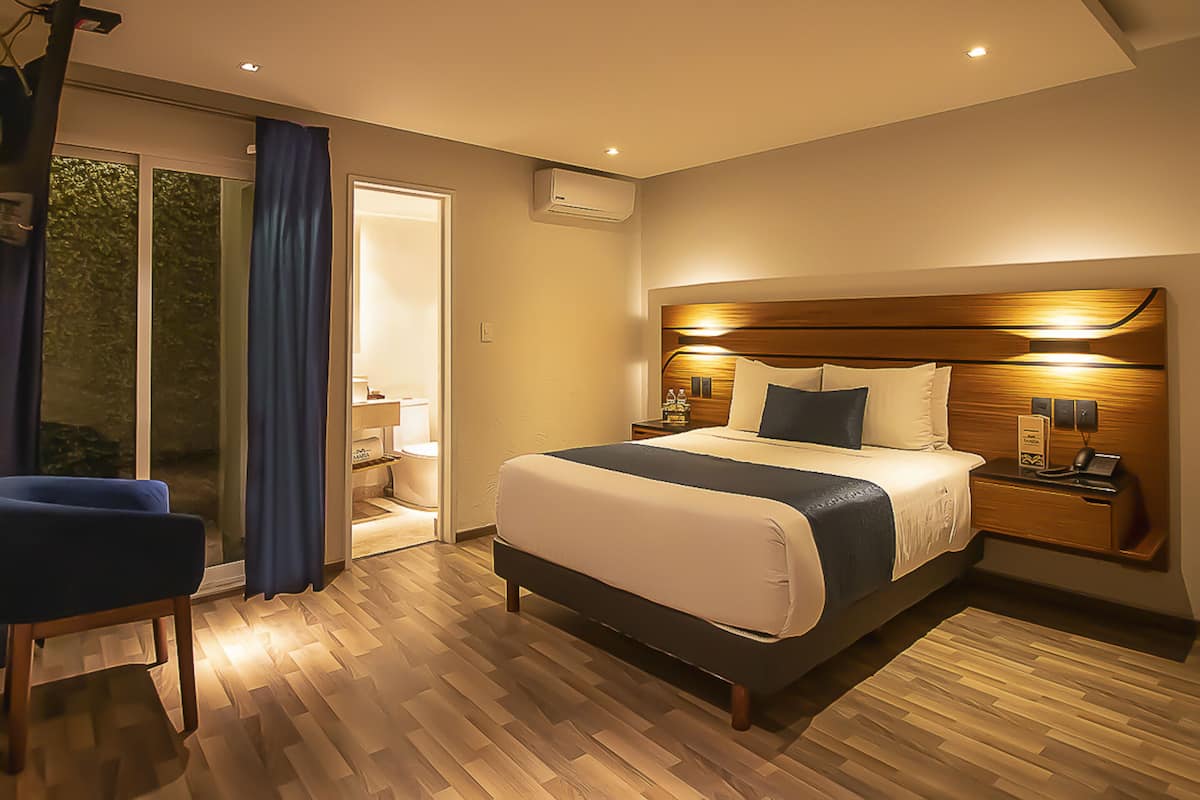 Een moderne hotelkamer met een groot bed, een nachtkastje met telefoon en lamp, een stoel en een glazen schuifdeur naar een badkamer.