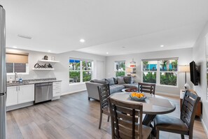 A modern kitchen with white cabinets, a dining area with a round table and chairs, and large windows offering a view of greenery. - ADA Compliance - 1 Bedroom/1 Bath Villa (Ground Level) - Unit 8 (Nokomis)