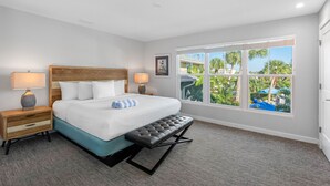 Habitación de hotel con una cama grande, un banco, una mesita de noche y una ventana con vistas a palmeras.