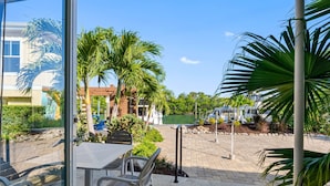 Area patio con tavolo e sedie, circondata da palme e vista su una piscina e edifici.