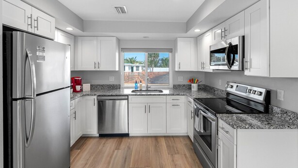 Una cocina moderna con electrodomésticos de acero inoxidable, armarios blancos, encimeras de granito y un ventanal con vistas a palmeras.
