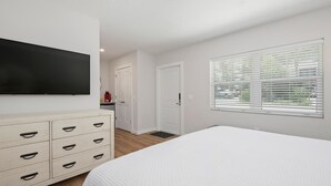 Une chambre avec un grand lit, un téléviseur fixé au mur, une commode avec des tiroirs et une fenêtre avec des stores.
