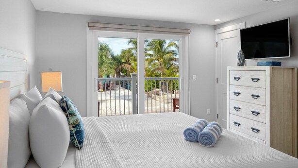 Ein Schlafzimmer mit Bett, Kissen, Fernseher, Kommode und einem Balkon mit Blick auf Palmen.