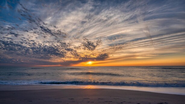Une plage où l’on voit des empreintes de pas menant à l’eau, un coucher de soleil et un ciel nuageux.