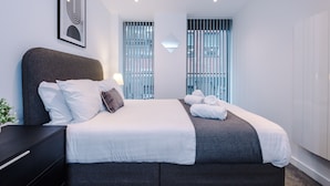 A modern hotel room with a large bed, a bedside table, a lamp, and a window with blinds. - Hilltop Serviced Apartments - Piccadilly (Manchester)