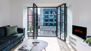 A modern living room with a sofa, a coffee table, a TV, and a balcony with a city view. - Hilltop Serviced Apartments - Piccadilly (Manchester)