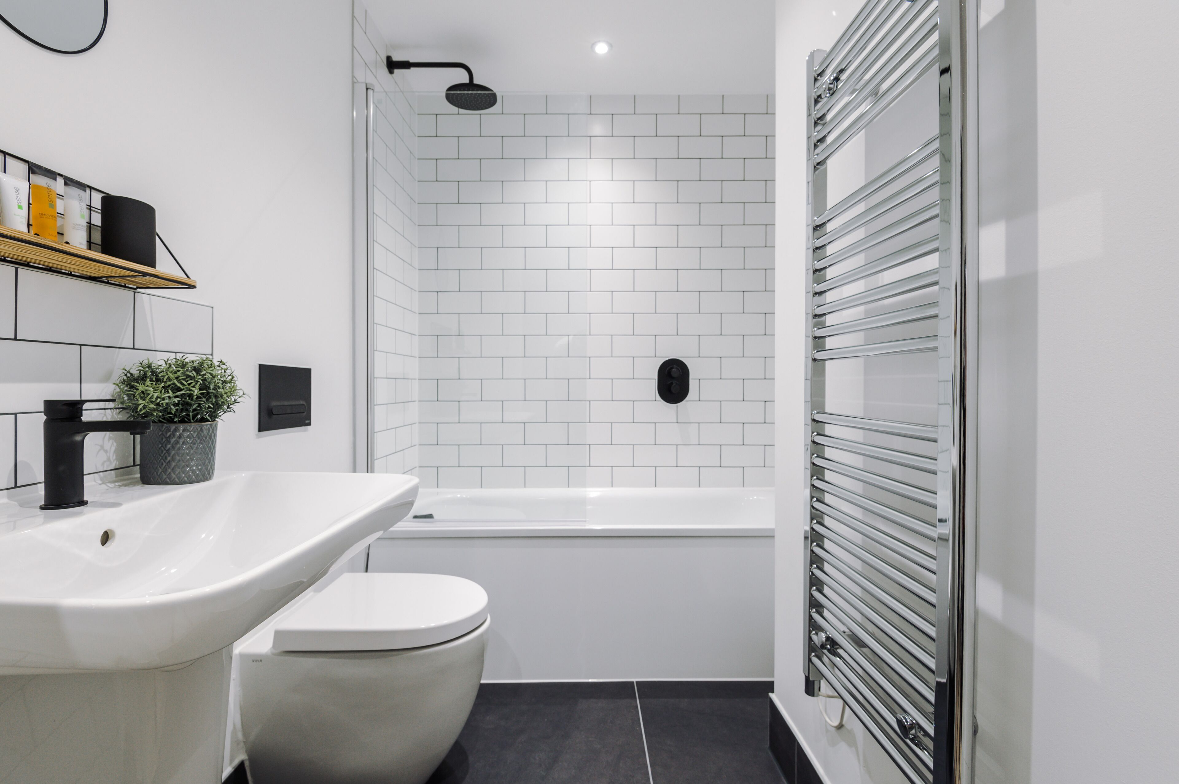 Ein modernes Badezimmer mit einem weißen Waschbecken, einer Toilette und einer Badewanne. Ein Regal mit Pflegeprodukten und ein Topf mit einer Pflanze sind vorhanden.
