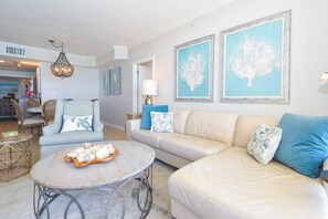 Un salon avec un canapé beige, une table basse ronde et un lustre. Deux œuvres d’art encadrées ornent le mur.