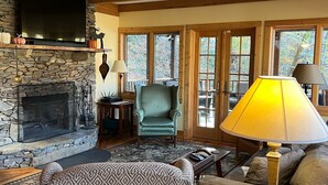 A cozy living room with a stone fireplace, a television mounted on the wall, and a comfortable seating area with a patterned armchair and a sofa. - 10 Star River Front Retreat - Water Sports, Hiking, Campfires by the River (Sparta)