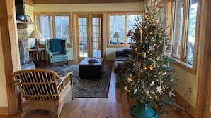 A cozy living room with a large Christmas tree, wooden furniture, and large windows. - 10 Star River Front Retreat - Water Sports, Hiking, Campfires by the River (Sparta)