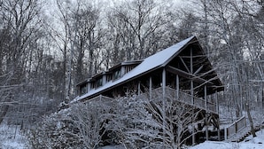A wooden cabin with a sloped roof and a balcony, surrounded by snow-covered trees and bushes. - 10 Star River Front Retreat - Water Sports, Hiking, Campfires by the River (Sparta)