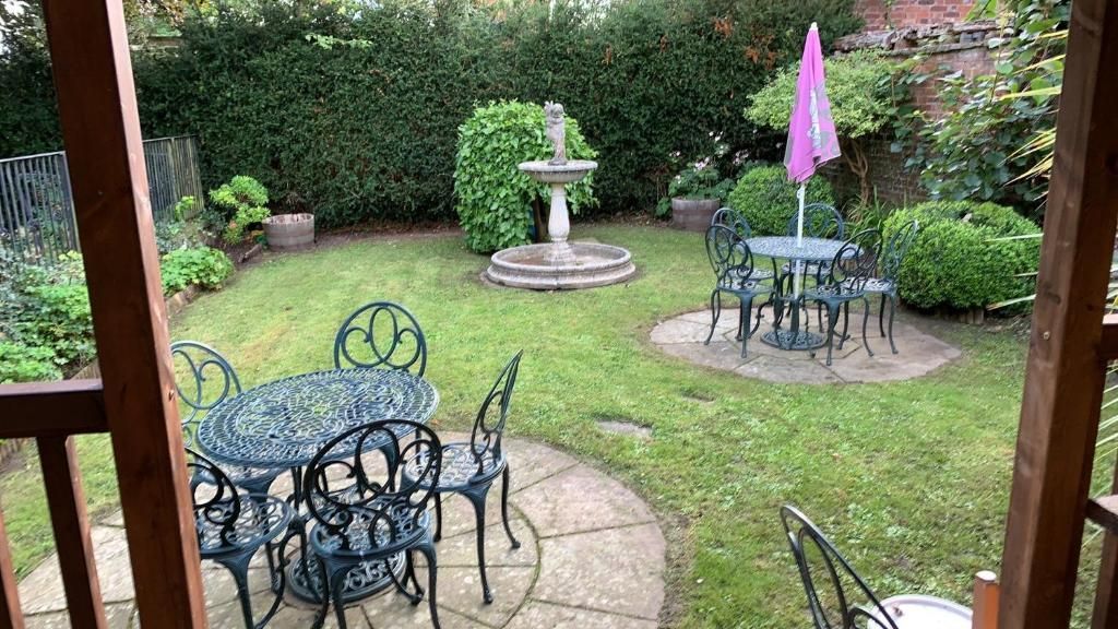 Eine Terrassenfläche mit rundem Tisch und Stühlen, umgeben von einem gepflegten Rasen und einer Zierbrunnen.