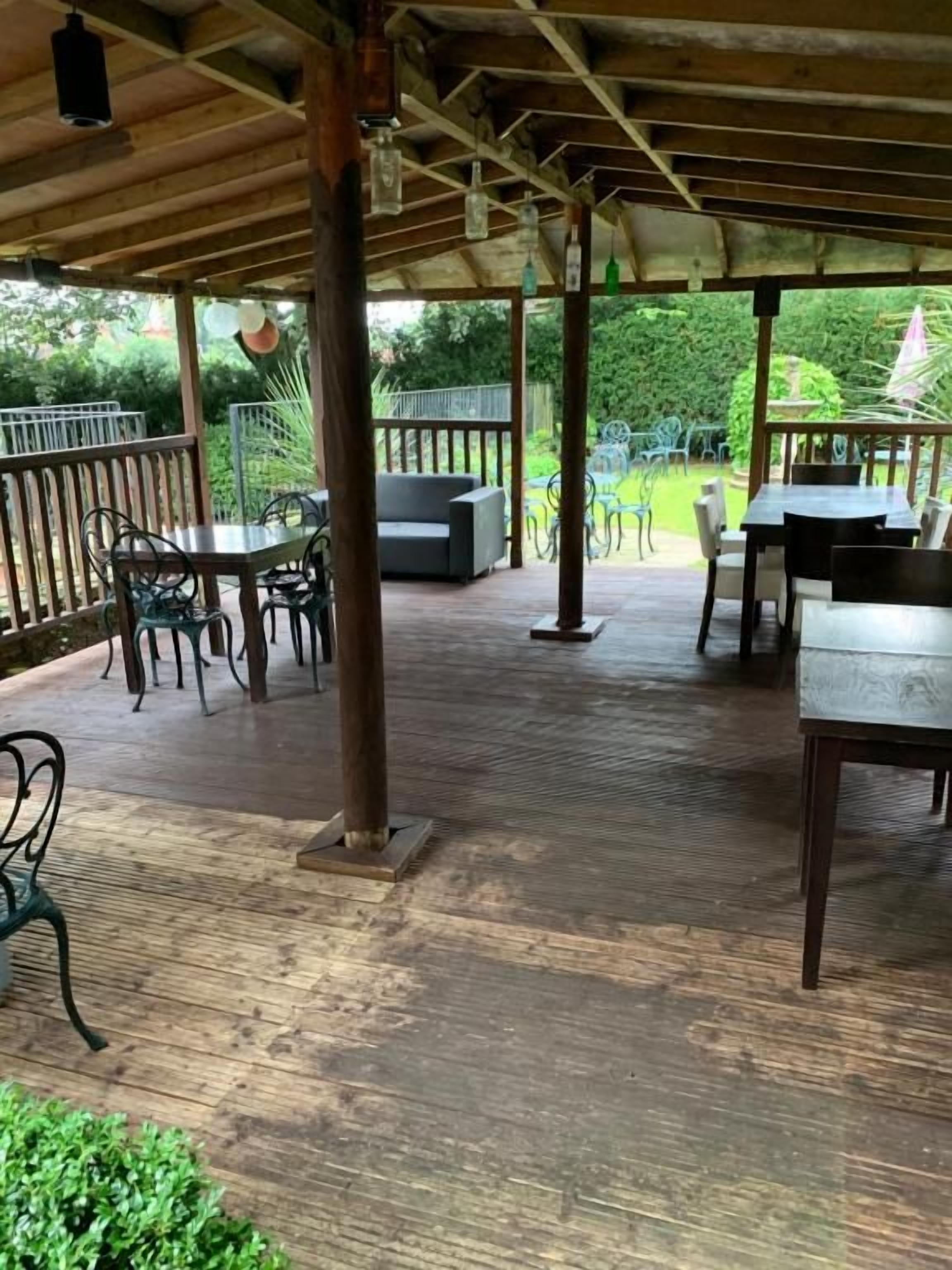 Un espace extérieur couvert, aménagé en patio, avec un plancher en bois, des rampes en métal et des coins salon comprenant des tables et des chaises.