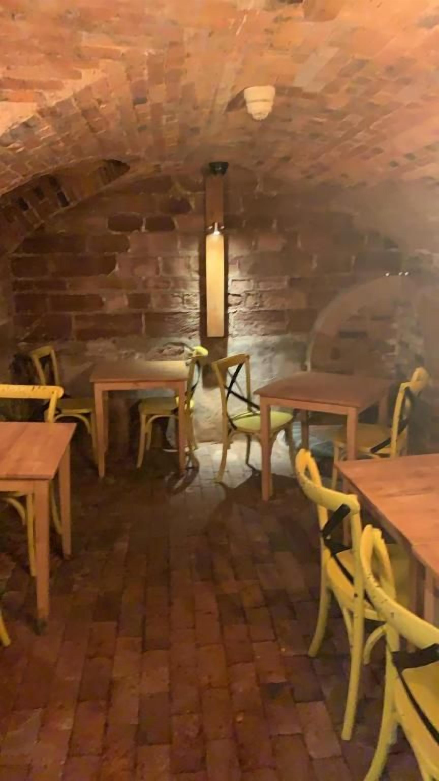 An underground dining area with brick walls, wooden tables and chairs, and a central light fixture.