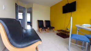 Una habitación con pared amarilla, un televisor montado en la pared, una mesa con una silla y dos sillones.