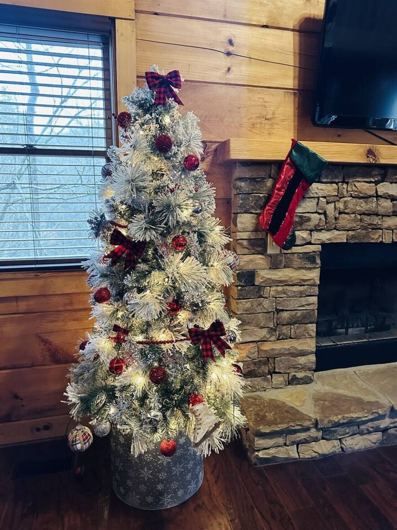 A Christmas tree with ornaments and a bow, placed in a room with a stone fireplace and a wooden wall.