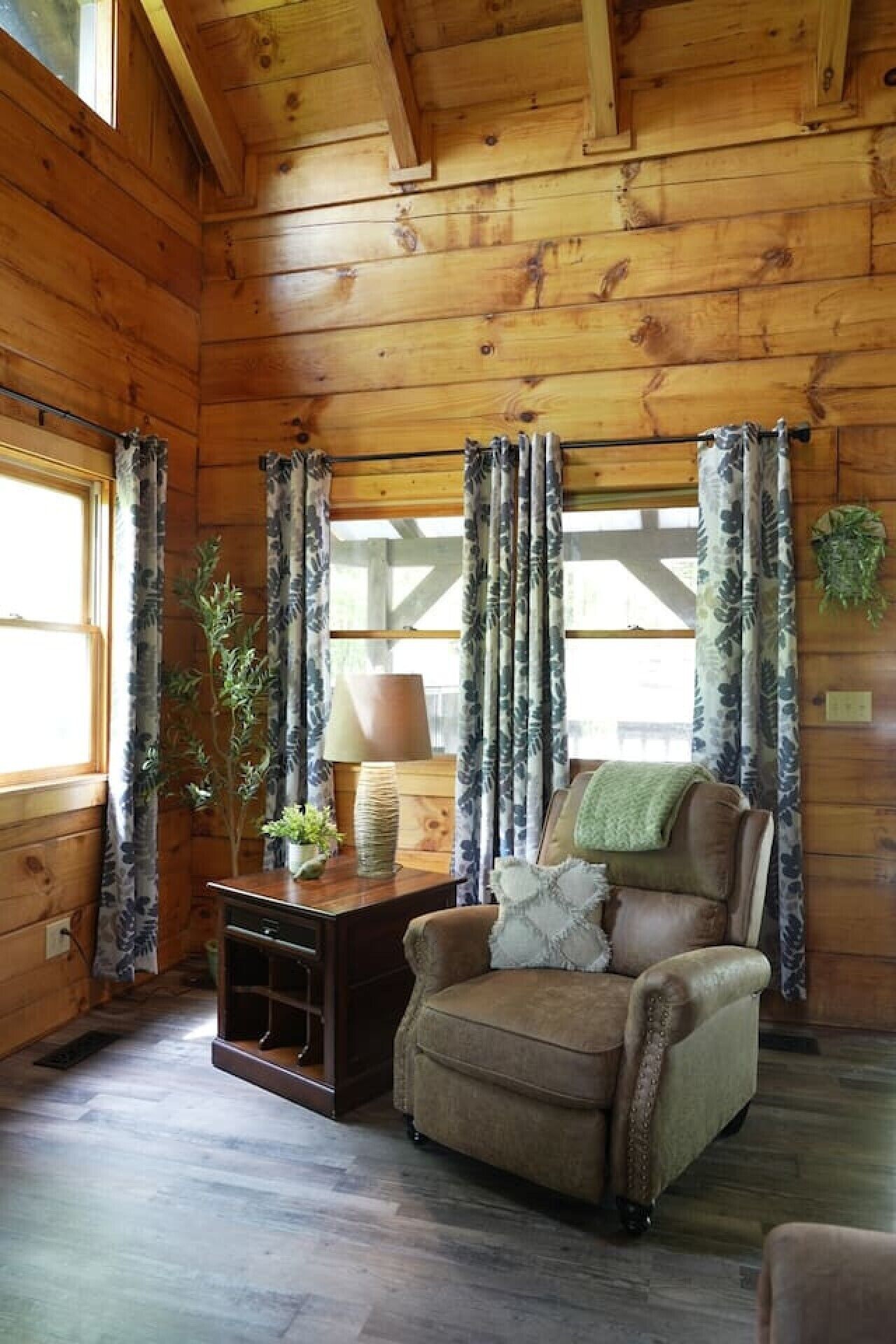 A cozy cabin interior with wooden walls, a leather armchair, a side table with a lamp, and curtains.
