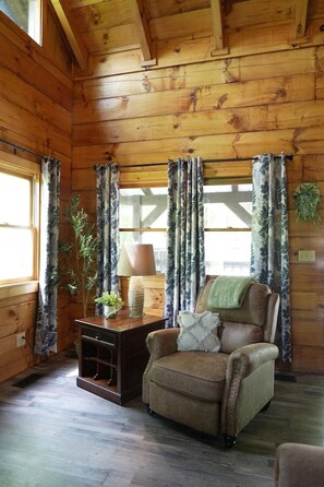 A cozy cabin interior with wooden walls, a leather armchair, a side table with a lamp, and curtains.