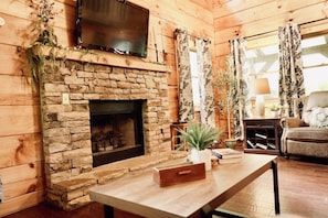 A cozy living room with a stone fireplace, a flat-screen TV mounted above it, and a comfortable seating area with a sofa and a table.