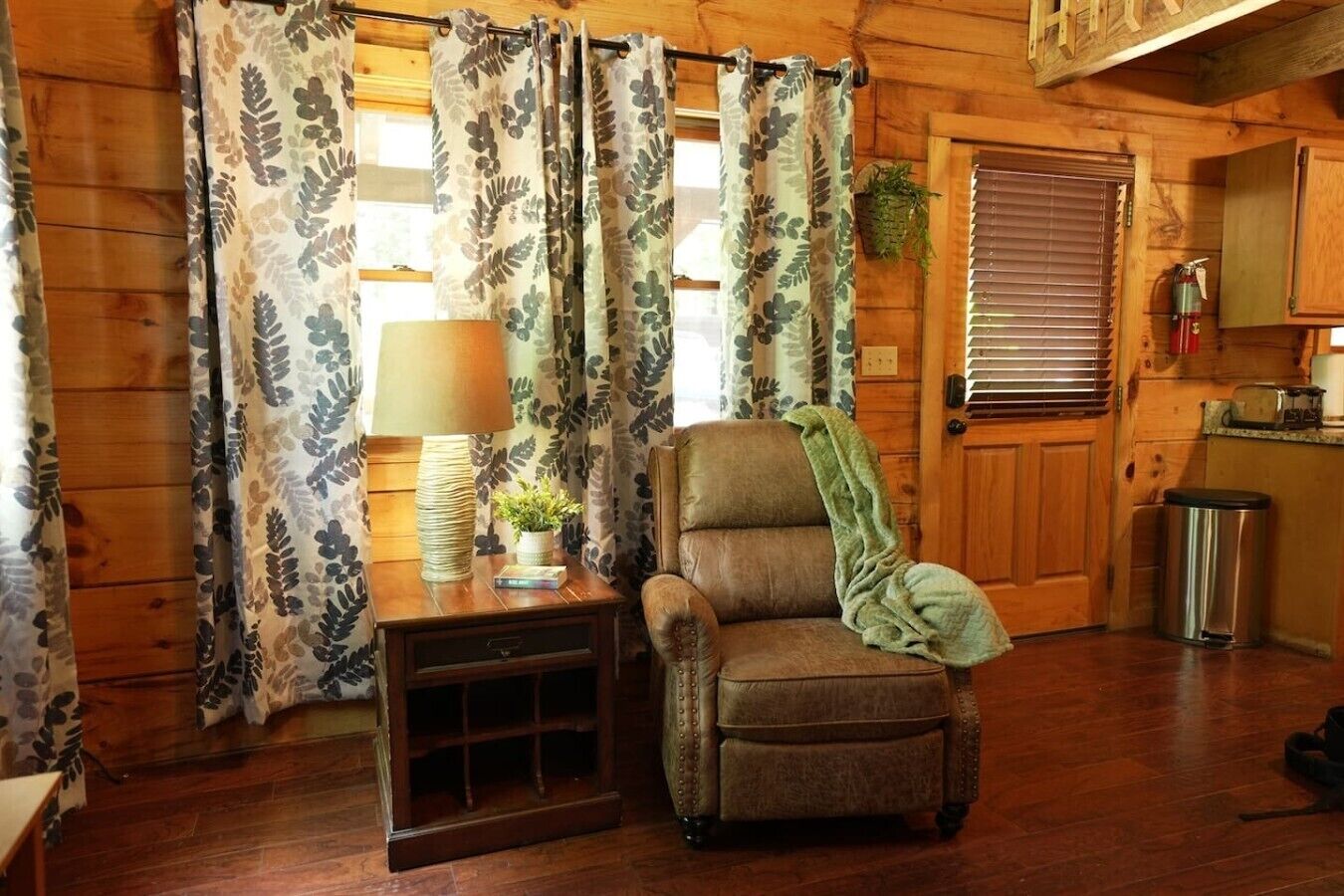 Een gezellig interieur van een vakantiehuisje met een houten vloer, een leren fauteuil, een bijzettafeltje met een lamp en gordijnen met een bladmotief.