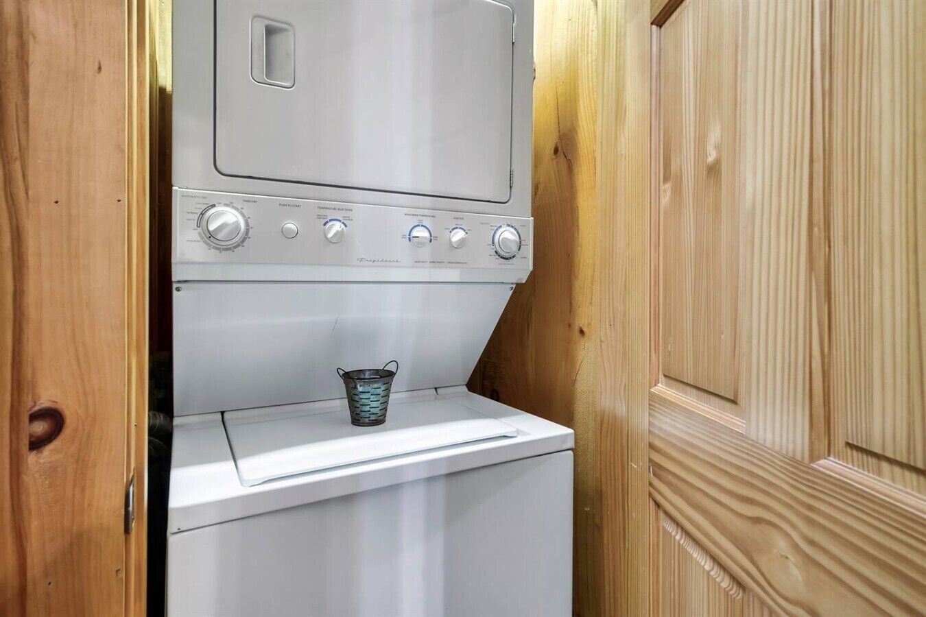 A compact laundry area with a washing machine and a drying machine, a metal bucket, and wooden paneling.