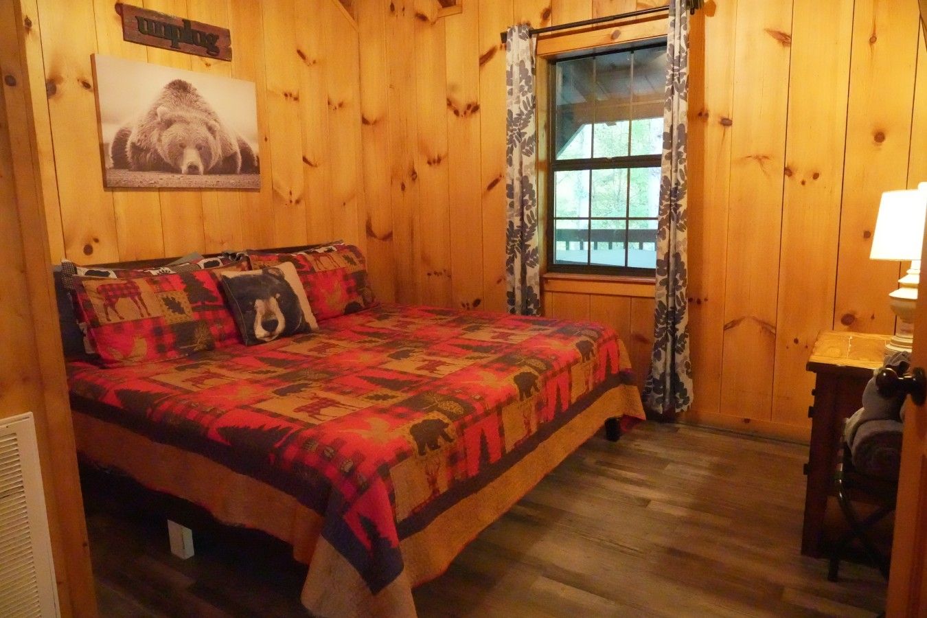 Une pièce en bois avec un lit, une fenêtre avec des rideaux, une lampe fixée au mur et un coussin à motifs d’ours.