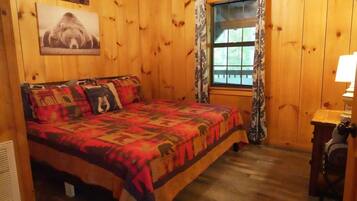 Une pièce en bois avec un lit, une fenêtre avec des rideaux, une lampe fixée au mur et un coussin à motifs d’ours.