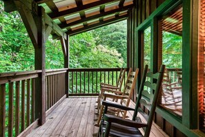 A wooden deck with a railing, wooden chairs, and a view of a forested area.