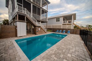 Une piscine avec une terrasse en bois, un espace couvert doté d’un escalier et une zone délimitée par une clôture, aménagée avec des chaises longues.