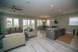 A spacious living room with a large window, a ceiling fan, and a dining area with a view of the outdoors. - Upscale Beach Front Home w/Elevator, Private Pool, Fire Pit! (Cape San Blas)