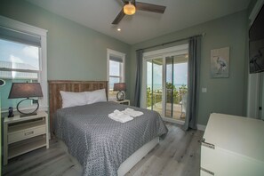A bedroom with a bed, bedside lamps, a ceiling fan, a large window with a view, and a sliding glass door leading to an outdoor area. - Upscale Beach Front Home w/Elevator, Private Pool, Fire Pit! (Cape San Blas)