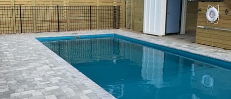 Ein rechteckiger Pool mit Fliesenveranda, umgeben von einem Holzzaun und einem Gebäude mit blauer Schrägverkleidung und weißen Geländern.