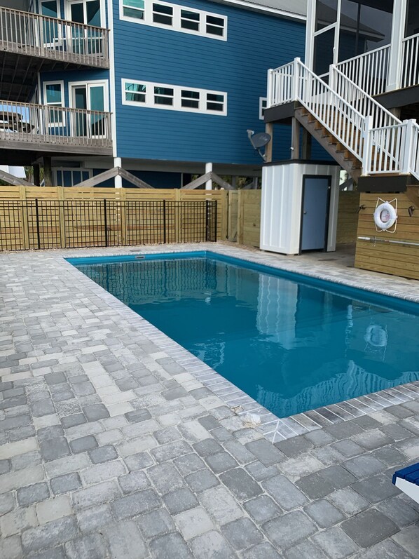 A rectangular swimming pool with a tiled deck, surrounded by a wooden fence and a building with blue siding and white railings. - Upscale Beach Front Home w/Elevator, Private Pool, Fire Pit! (Cape San Blas)